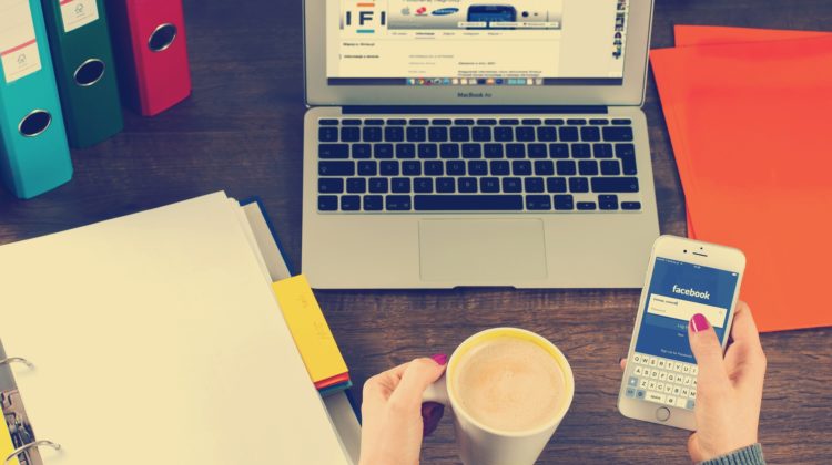 A woman holding a coffee cup while browsing Facebook in front of her laptop