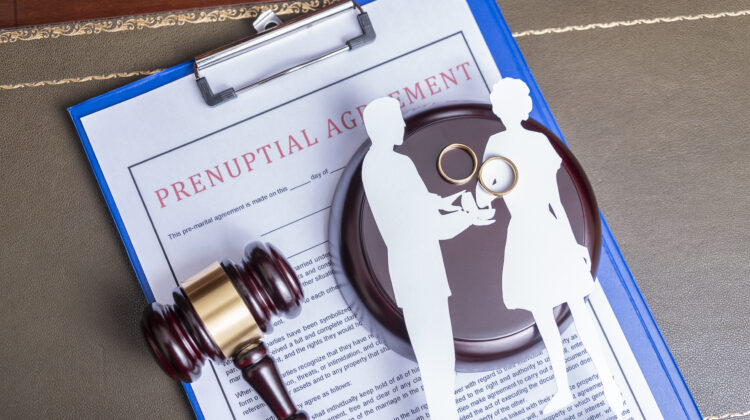 Prenuptial agreement document on a clipboard with a gavel and wedding rings on top of it.