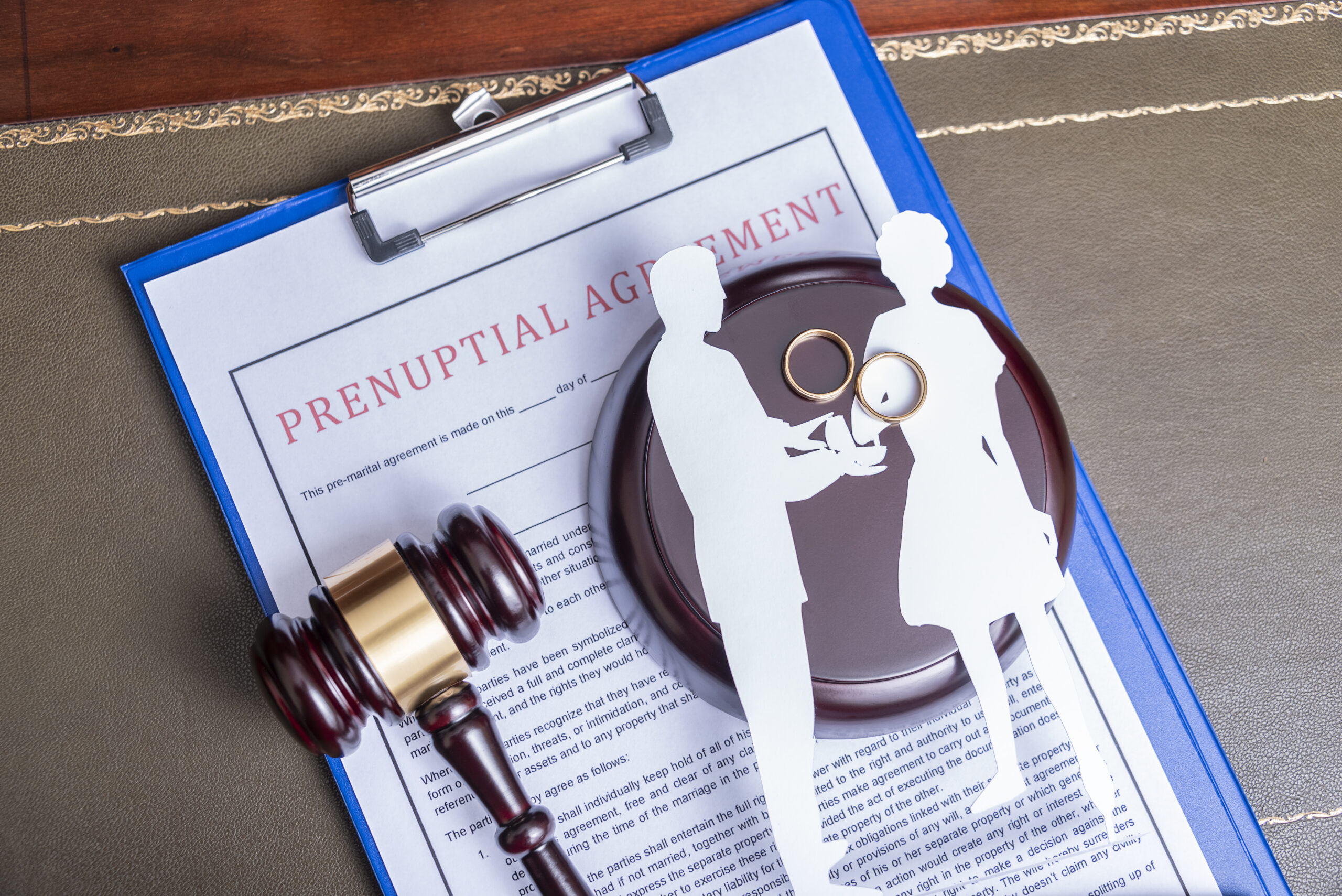 Prenuptial agreement document on a clipboard with a gavel and wedding rings on top of it.